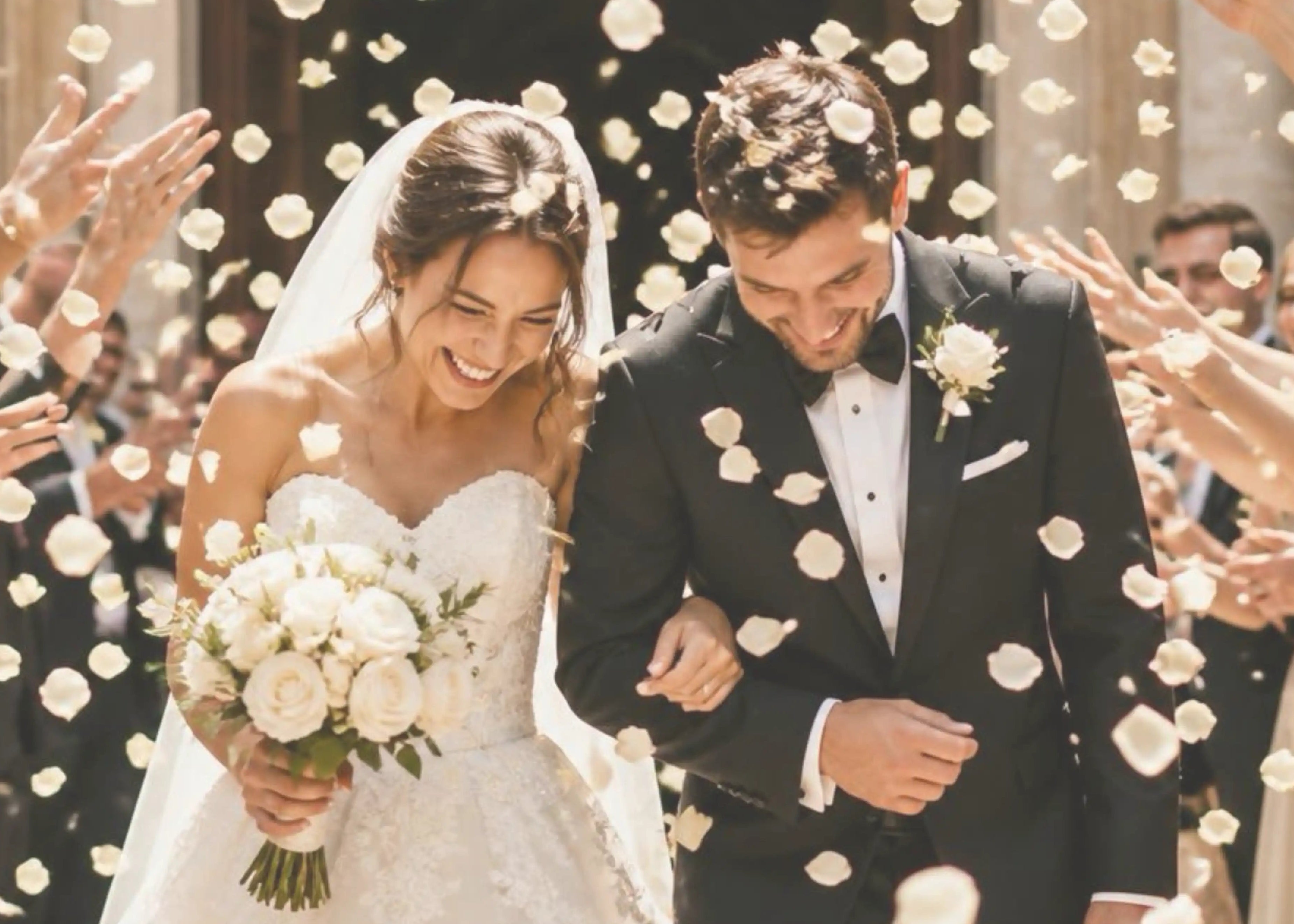 Wedding couple walking through a shower of white flowers with a crowd of people in the background.