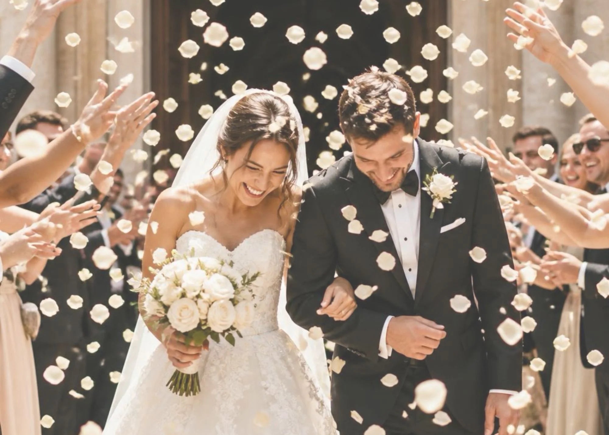 Wedding couple walking through a shower of white flowers with a crowd of people in the background.